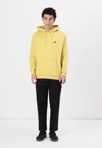 Felpa gialla con cappuccio, dotata di tasca anteriore e logo, abbinata a pantaloni neri e sneakers nere. Design semplice e vestibilità rilassata.