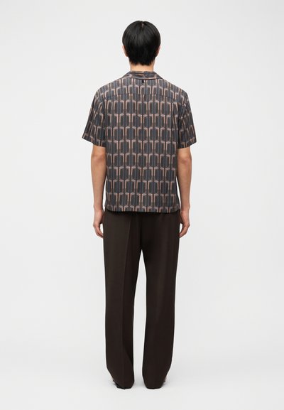Moški, obrnjen stran, oblečen v kratkoročno srajco z vzorcem geometrijskih oblik in ohlapne temne hlače na enostavni podlagi.