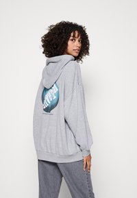 Sudadera gris oversize hecha de una tela suave, con un estampado gráfico en teal y blanco en la parte posterior, con capucha ajustable y puños acanalados.