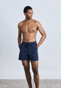 Μπλε ναυτικό μαγιό με ελαστική μέση, κλείσιμο με κορδόνι και πλαϊνές τσέπες. Διαθέτει διακριτό σχέδιο υφής και λογότυπο.