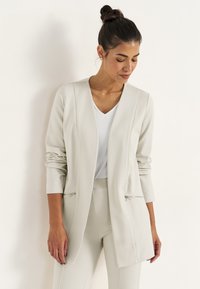 Ljust beige skräddarsydd jacka med långa ärmar, öppen fram och dekorativa siddragkedjor. Slätt tyg med en svag lyster och strukturerad design.