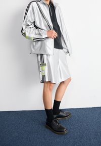 Person som bär en silver Adidas-träningsjacka och shorts med svarta ränder, svarta strumpor, svarta skor, står på en blå matta mot en vit vägg.