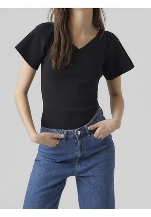 Vrouw draagt een nauwsluitend zwart V-hals topje met korte mouwen en een high-waisted blauwe spijkerbroek, handen in de voorzakken, grijze achtergrond.