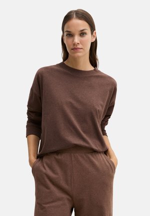 Frau mit glattem braunen Haar, die ein lockeres braunes Langarmshirt und passende Hose trägt, mit den Händen in den Taschen vor einem einfarbigen Hintergrund.