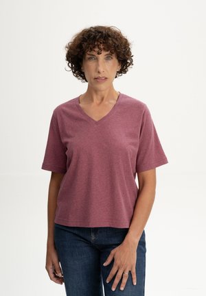 Frau mit kurzen lockigen Haaren, die ein mauvefarbenes V-Ausschnitt-T-Shirt und dunkelblaue Jeans trägt, vor einem schlichten hellen Hintergrund stehend.