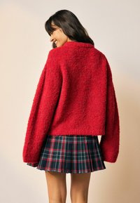 Rød, fluffy, cropped sweater med brede ærmer, parret med en sort og rød ternet, plisseret nederdel, med synlig søm og plisseringer i bunden.