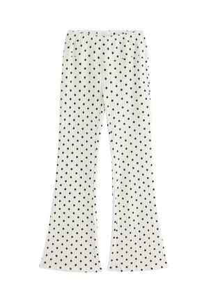 Jeune femme debout, portant un haut noir sans manches, un pantalon blanc à pois noirs et des chaussures noires, sur un fond clair uni.