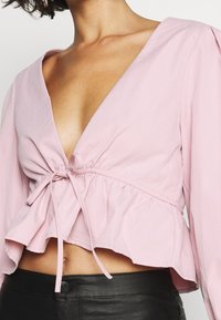 Blouse rose à manches longues avec un décolleté en V profond et un détail de nœud froncé à la taille, présentant un ourlet volanté et une texture lisse.
