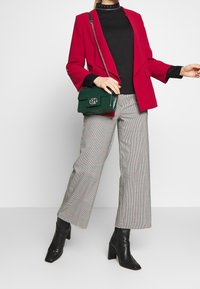 Röd blazer med asymmetrisk design, svart polotröja, rutiga vida byxor och en svart och grön texturerad handväska.