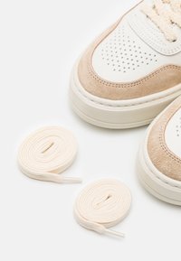 Baskets beige et blanches avec des détails perforés et deux paires de lacets beiges roulés sur un fond blanc.
