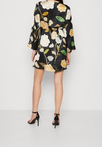 Robe noire à fleurs avec de grands motifs de roses blanches et jaunes, taille cintrée et manches évasées. Portée avec des sandales à talons hauts noires.