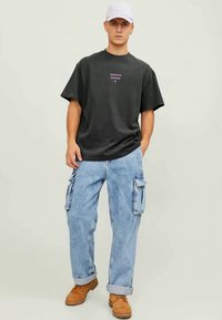 Jovem vestido com um boné de basebol branco, uma t-shirt preta "Premium Studios", jeans cargo azul claro e botas de trabalho castanhas, posando em frente a um fundo simples.