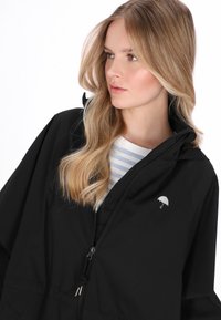 Fekete vízálló kabát kapucnival és ezüst esernyő logóval a mellkason. A modell egy világos csíkos felsőt visel alatta.