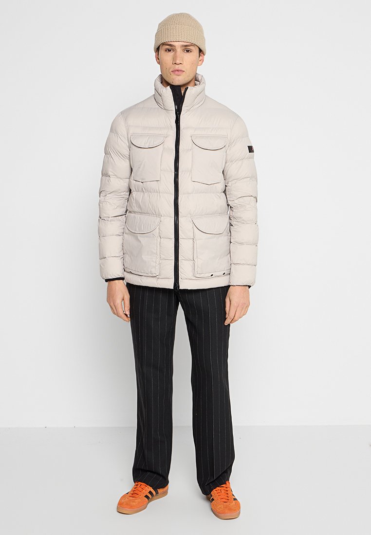 Giacca a vento beige chiaro con zip nera a contrasto, quattro tasche frontali e colletto alto. Indossata con pantaloni a righe e scarpe arancioni.