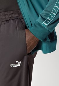 Puma sporta tērps, kurā ietilpst zila jaka ar rāvējslēdzēju un logo akcentiem, kā arī melnas bikses ar baltu logo, kas izgatavotas no gludas, vieglas auduma.