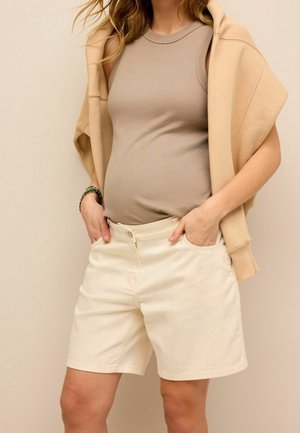 Femme portant un haut beige sans manches et un short crème aux genoux, avec un sweat-shirt marron clair drapé sur les épaules.