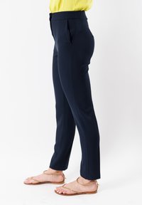 Pantaloni navy blue su misura con pieghe frontali e tasche laterali, realizzati in tessuto liscio, abbinati a sandali piatti dorati luccicanti.