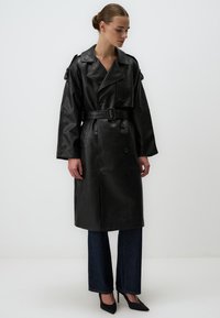 Trench coat in pelle nera con maniche ampie, bottoni doppi e vita con cinturino. Indossato sopra pantaloni in denim scuri e tacchi neri.