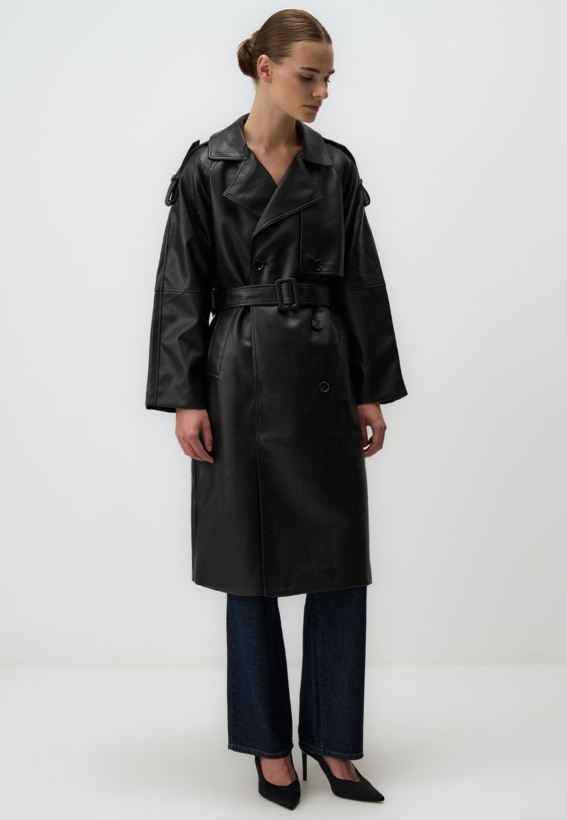 Trench coat in pelle nera con maniche ampie, bottoni doppi e vita con cinturino. Indossato sopra pantaloni in denim scuri e tacchi neri.