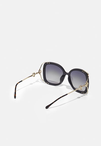 Lunettes de soleil surdimensionnées avec des montures noires, des verres dégradés et des branches accentuées en or. Détails texturés sur les bords de la monture et les branches.