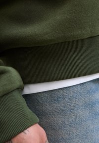 Närbild av en person som bär en grön ribbad tröja över en vit skjorta och ljusblå denimjeans, vilket visar en tatuerad hand.