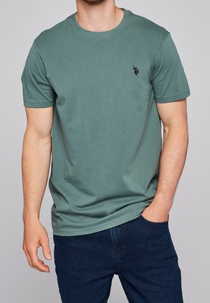 Groene katoenen T-shirt met korte mouwen, ronde hals en een klein zwart logo op de borst. Draagt met donkerblauwe jeans.