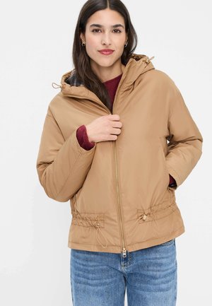 Frau trägt eine hellbraune, kapuzenbedeckte Winterjacke über einem burgunderroten Pullover und blauen Jeans, mit einer Hand, die gerade den Reißverschluss der Jacke hochzieht.