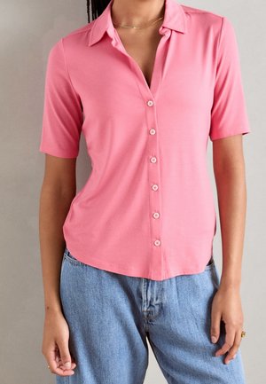 Vrouw draagt een roze korte mouwen blouse met kraag en lichtblauwe spijkerbroek, staand tegen een effen lichtgrijze achtergrond.