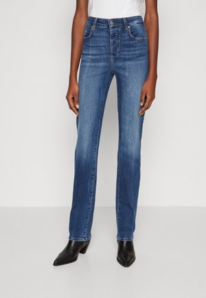 Femme portant un jean bleu taille mi-haute droit et des bottines à talons noires à bout pointu sur un fond clair.
