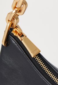 Black leather handbag corner with a gold zipper pull and chain detail. Features a gold zip and fabric accent with branding on the hardware.