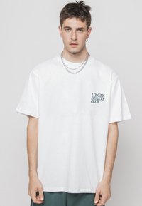 Balts kokvilnas T-krekls ar brīvu piegriezumu, ar nelielu zaļu "Lonely Hearts Club" druku kreisajā krūtīs un īsām piedurknēm.