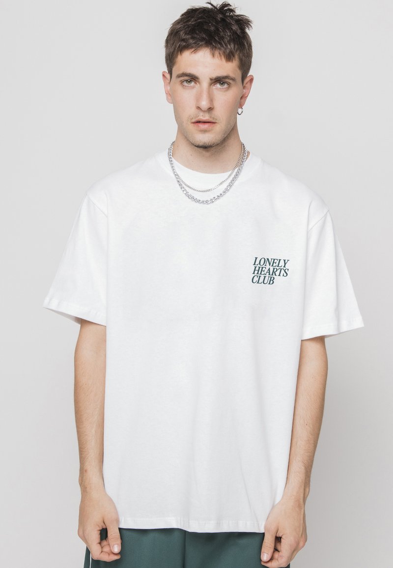 Balts kokvilnas T-krekls ar brīvu piegriezumu, ar nelielu zaļu "Lonely Hearts Club" druku kreisajā krūtīs un īsām piedurknēm.