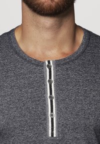 Tummanharmaa henley-paita, jossa on pyöreä kaula-aukko, viiden napin avaus musta-valkoisilla raidoilla varustettuna ja tekstuurikangas.