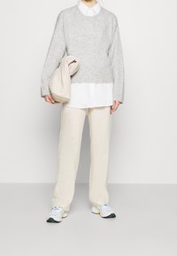 Persona in pantaloni beige, sneakers bianchi, camicia bianca a strati sotto un maglione grigio, che tiene una clutch beige testurizzata davanti a uno sfondo neutro.