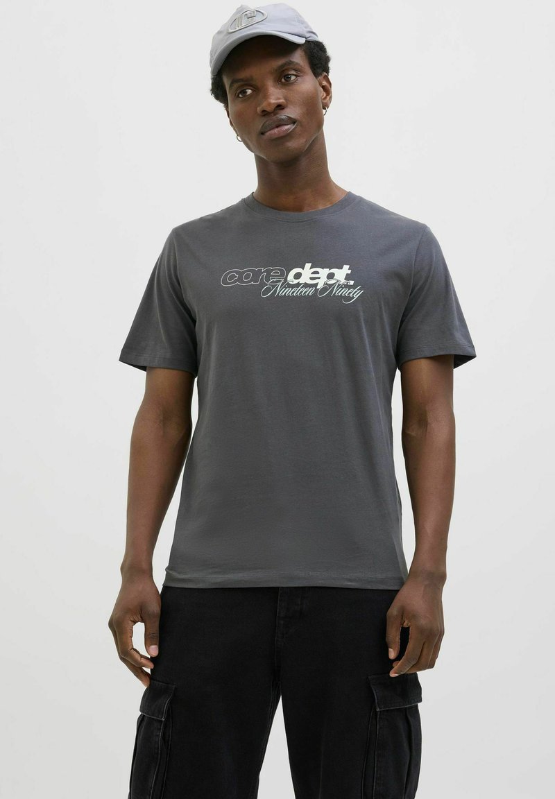 Grå bomulds T-shirt med korte ærmer og rund halsudskæring. Forsiden har trykt hvid tekst; iført sorte cargo bukser.