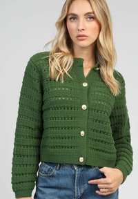 Cardigan en tricot vert avec des motifs de rayures horizontales, des poignets et un ourlet côtelés, et cinq grands boutons ronds sur le devant.