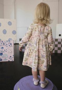 Blomstret børnekjole i pastelrosa med lange ærmer, lagdelt design, enhjørninger og regnbuer, kombineret med hvide sko og blonder sokker.