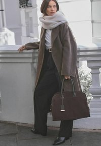 Bruine wollen jas over een witte top, zwarte high-waisted broek. Houdt een donker suède handtas met leren accenten en een rechthoekig label vast.