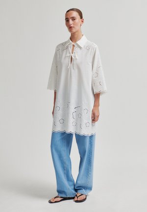 Frau steht und trägt eine weiße Tunika mit Lochstickerei und Blumenmuster, dreiviertellangen Ärmeln, hellblauen weiten Jeans und schwarzen Flip-Flops vor einem neutralen Hintergrund.