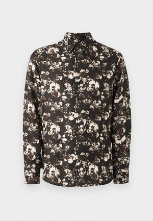 Mand iført sort-hvid blomstret langærmet skjorte, brune bukser og hvide sneakers, står med den ene hånd i lommen og kigger mod højre.