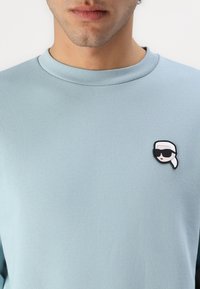 Ανοιχτό μπλε πουλόβερ με στρογγυλή λαιμόκοψη, διαθέτοντας μία μαύρη και λευκή παραλλαγή μιας φιγούρας με γυαλιά ηλίου στην αριστερή περιοχή του στήθους.