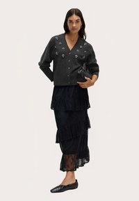 Cardigan gris foncé avec des embellissements floraux, jupe noire superposée avec bordure en dentelle, et chaussures plates noires, complétés par un petit sac à main.