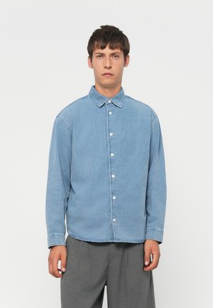 Man wearing light blue denim button-up shirt with long sleeves and gray pants, standing against a plain white background.