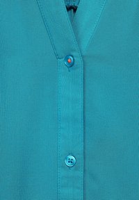 Chemise turquoise à boutons, avec un design en col V, texture lisse, et un bouton rond orné d'une étoile rouge contrastante.