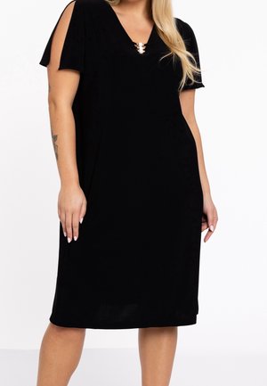 Kobieta w czarnej sukience do kolan z rozcięciem, krótkimi rękawami i perłowym zdobieniem przy dekolcie w kształcie litery V, stojąca na białym tle.