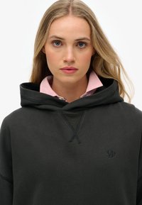 Sudadera negra con corte holgado, capucha con cordón y detalle cosido en el escote. Presenta un pequeño logo en el pecho y una camisa blanca con cuello rosa debajo.