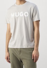 Muž oblečený v světle šedém tričku s nápisem "HUGO" bílými písmeny a olivově zelených kalhotách, zobrazený od ramen po boky.