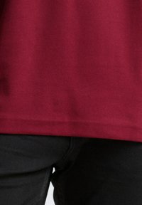 Bordeauxfarbenes Hemd, das über schwarzen Hosen mit sichtbaren Nähten und Stofftextur hängt.