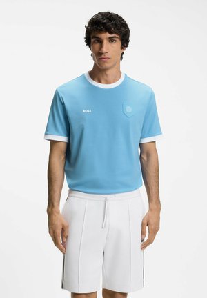 Jonge man draagt een lichtblauw T-shirt met witte kraag en manchetten, en witte short met trekkoord, staand tegen een witte achtergrond.