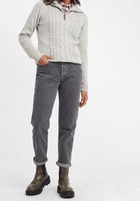 Pullover in maglia leggera grigia con zip a metà e tirazip in pelle, abbinato a jeans grigi scuri e stivali marroni alla caviglia.